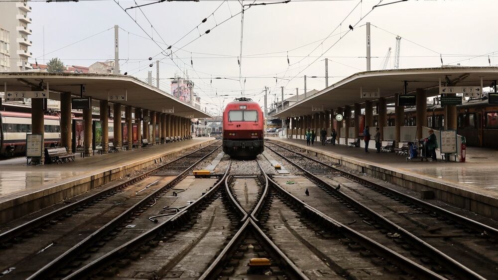 Estação de comboios com comboio vermelho ao centro, carris em primeiro plano