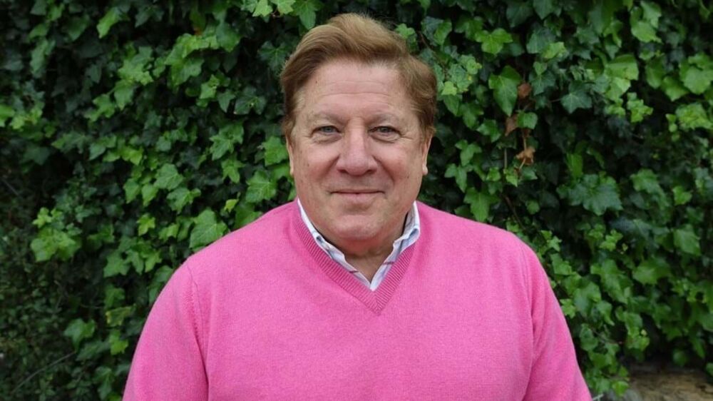 Retrato de homem sorridente com camisola rosa, em frente a uma parede de hera