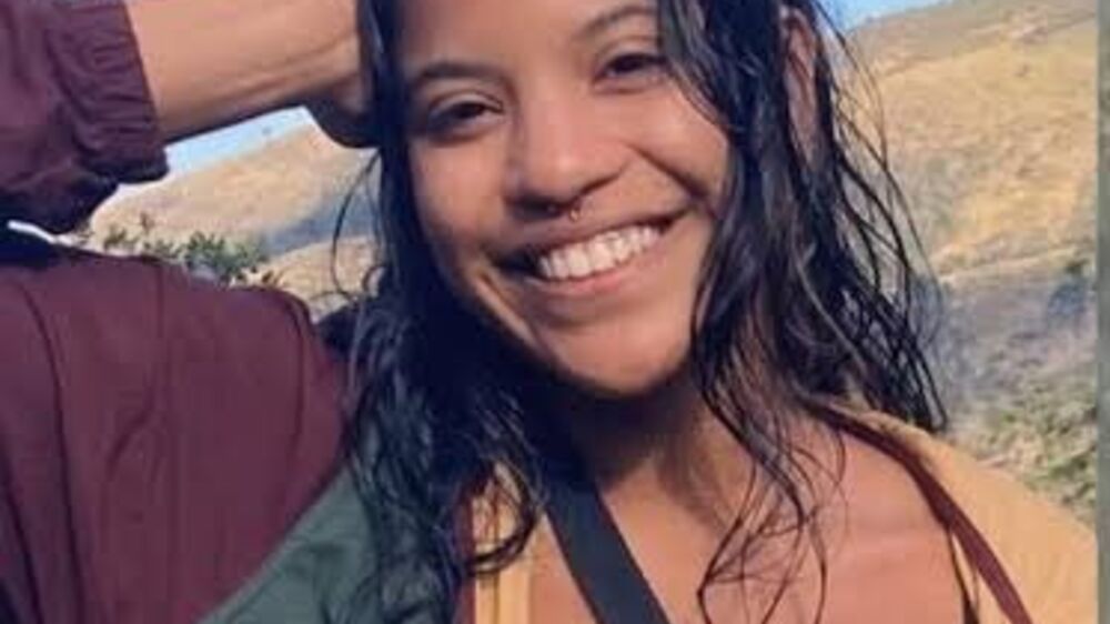 Mulher sorri com jaqueta e piercing no nariz, em cenário montanhoso