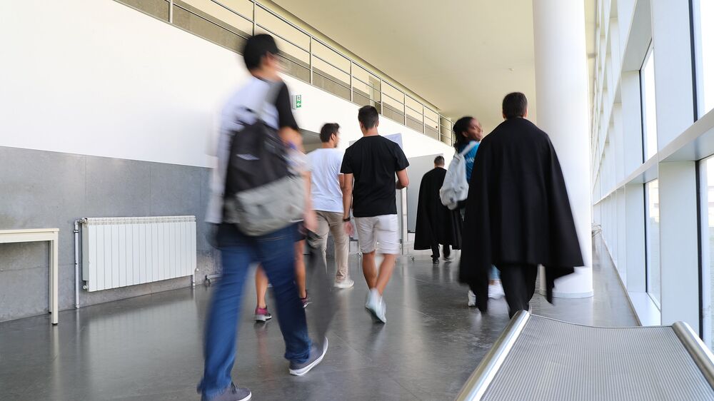Pessoas caminham num corredor com luz natural, algumas de capa preta