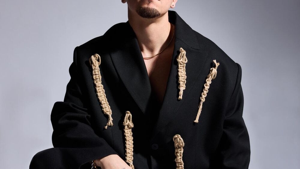 Homem posa com blazer preto com detalhes de corda bege