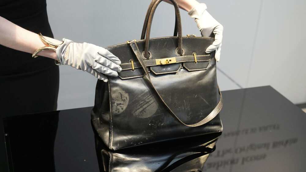 Mãos com luvas exibem mala de couro preta com detalhes dourados