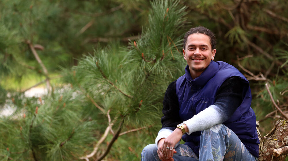 Homem sorri perto de pinheiro com colete azul e calças florais
